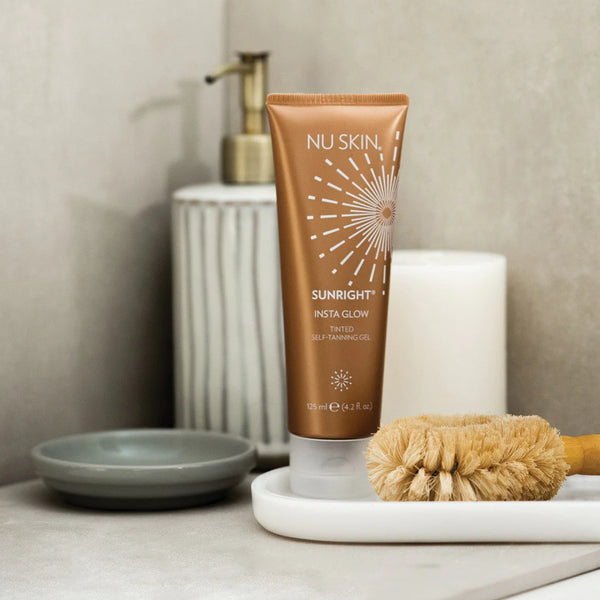 A tube of Sunright Insta Glow Tinted Self Tanning Gel by NuSkin, placed on a white tray with a brush, next to a candle and a bathroom sink.