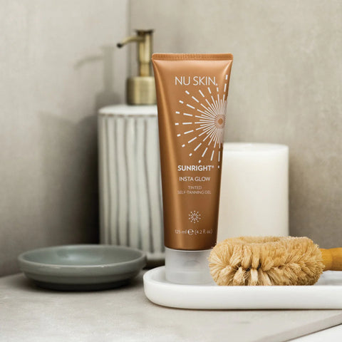 A tube of Sunright Insta Glow Tinted Self Tanning Gel by NuSkin, placed on a white tray with a brush, next to a candle and a bathroom sink.